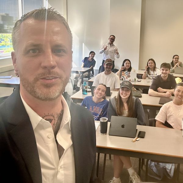 Jamie Gerrard taking a selfie in a classroom at Saint Mary's University, with smiling MBA students seated at desks with laptops behind him.