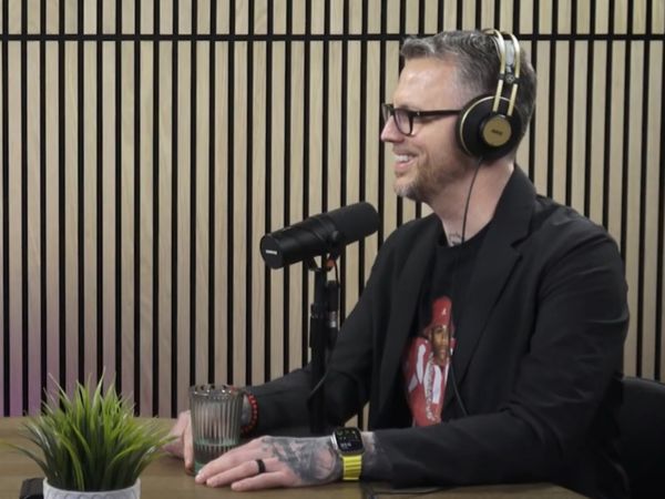 Jamie Gerrard smiling in headphones at a podcast microphone, wearing a black blazer over an LL Cool J T-shirt, in front of a wooden slat wall.