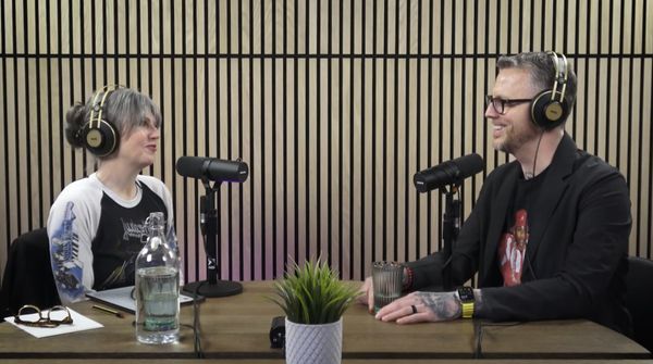 Carrie Forbes and Jamie Gerrard seated across a table in headphones, talking into microphones in front of a wooden slat wall.