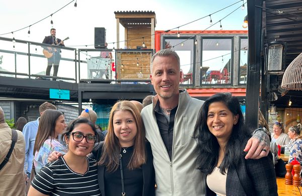 Jamie Gerrard and three others smiling arm in arm on a busy outdoor patio, with a musician playing guitar on the deck behind them.
