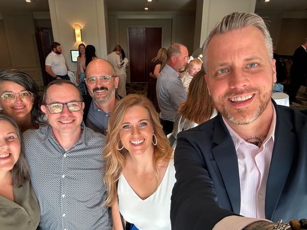 Jamie Gerrard and five smiling friends and colleagues gathered close for a group selfie at an indoor event.