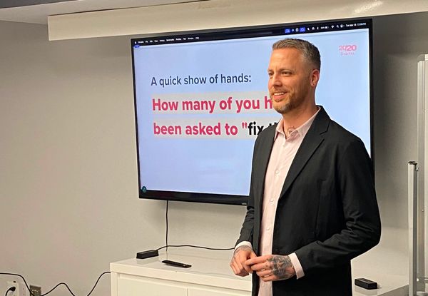 Jamie Gerrard beside a screen with a slide that reads: A quick show of hands: How many of you have been asked to fix the UX, partly hidden behind him.