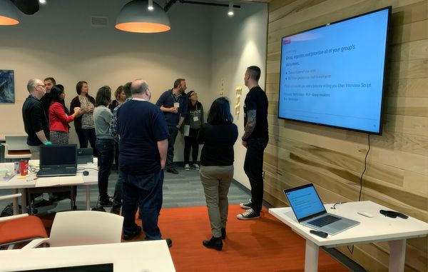Workshop participants and Jamie Gerrard gathered at a large screen with the instruction to group, organize, and prioritize their sticky notes.