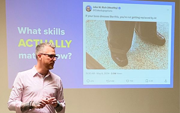 Jamie Gerrard presenting in front of a slide that asks, What skills actually matter now?, beside a projected social media post that reads, if your boss dresses like this, you're not getting replaced by AI, above a photo of dress pants and leather shoes.