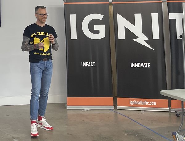 Jamie Gerrard presenting at IGNITE Atlantic in a Wu-Tang Clan T-shirt, beside IGNITE banners reading Impact and Innovate.