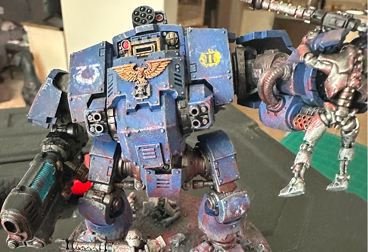 A blue Ultramarines Dreadnought with a gold winged skull, standing over battlefield rubble, holding a Necron Warrior in its left hand.